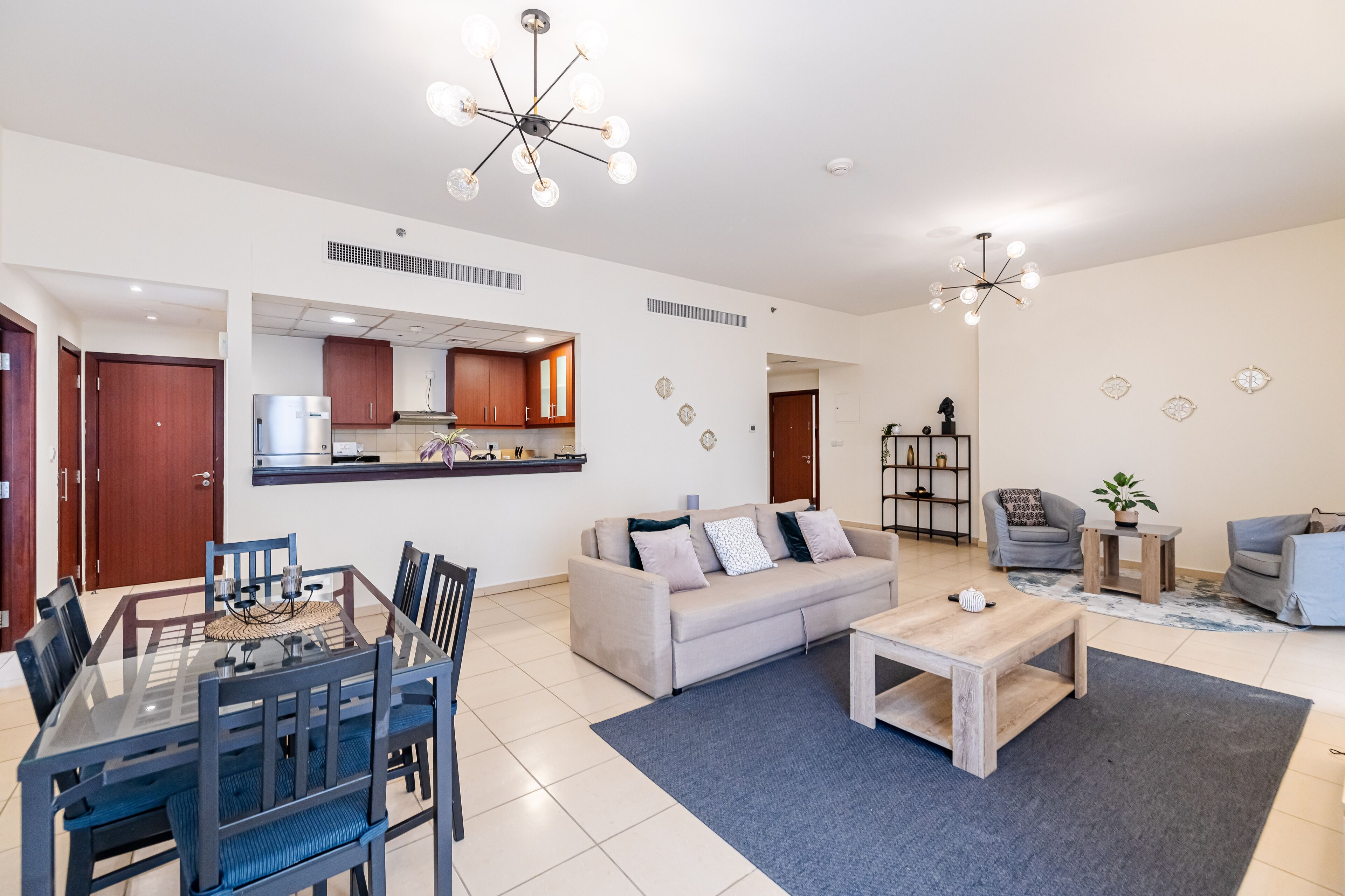 A modern living room with a sofa, coffee table, and dining area. There is a kitchen in the background with cabinets and a refrigerator.