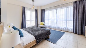 Moderni hotellihuone, jossa on suuri sänky, työpöytä ja tuoli. Huoneessa on suuret ikkunat, joissa on verhot, sekä lamppu ja matto.