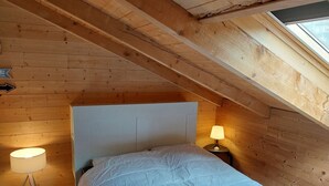 Ein kleines, gemütliches Schlafzimmer im Dachgeschoss mit Holzwänden und schrägen Decken. Es gibt ein Bett mit weißem Kopfteil, zwei Nachttischlampen und einen kleinen Teppich.