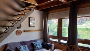 Ein gemütliches Zimmer mit einer Holztreppe, einer Couch mit Kissen, einem Korbsessel, einem kleinen Tisch und einem Fenster mit Blick auf Grünflächen und Gebäude.
