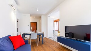 A modern living room with a blue sofa, a red pillow, a dining area with a table and chairs, and a large flat-screen TV mounted on a blue cabinet.