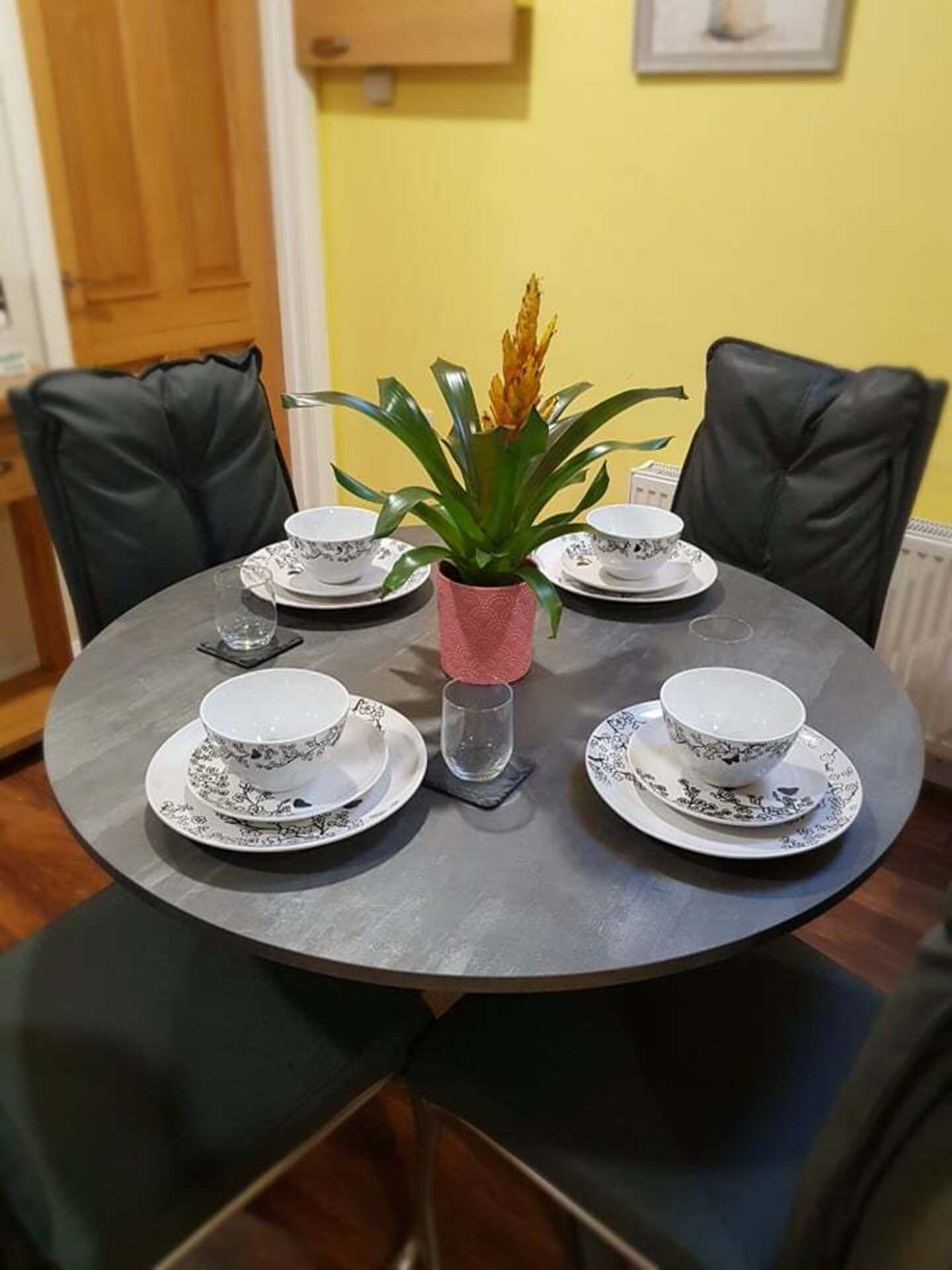 A round dining table with four white cups and saucers, a pink vase with a plant, and black leather chairs.
