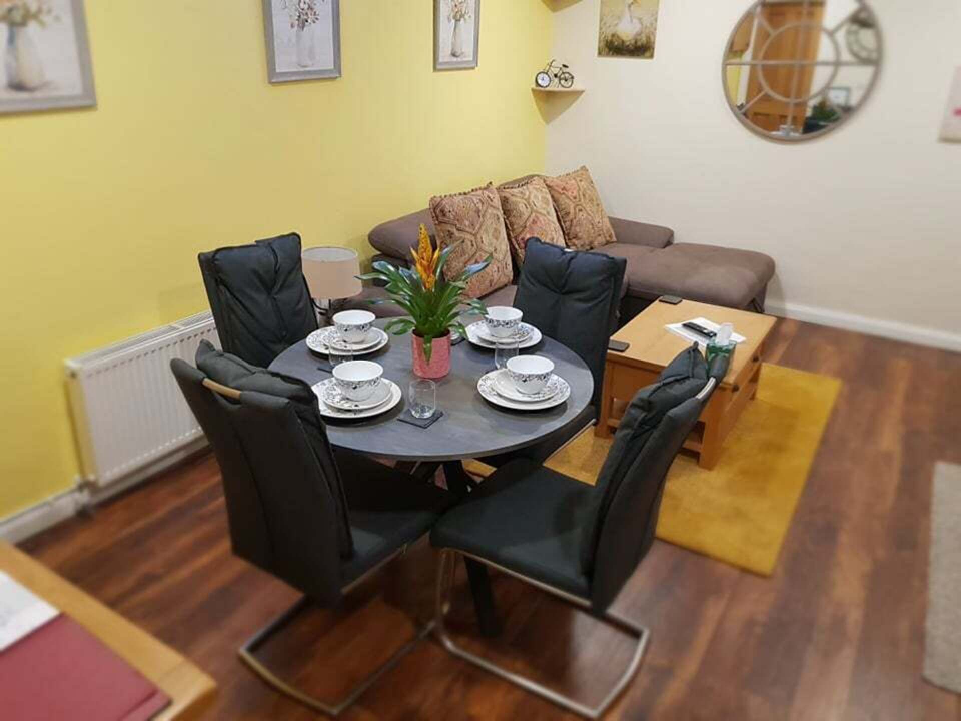 A dining area with a round table set for four, surrounded by black chairs. There is a sofa with patterned cushions in the background, a small wooden side table, and a mirror on the wall.