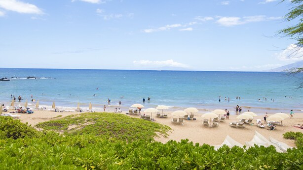 Une plage avec des chaises longues et des parasols, une eau d’un bleu limpide et une montagne qui se profile à l’horizon.