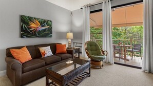 Un salon avec un canapé marron, une table basse en verre, un fauteuil en osier et un tableau représentant un oiseau du paradis.
