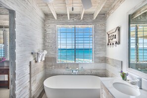 A bathroom with a large bathtub, a window with a view of the sea, and a wooden ceiling. - Charming Beachfront Villa - Sunset Reach (Mullins Beach)