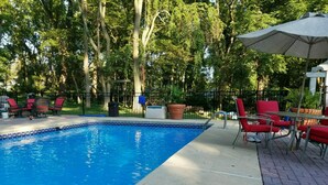 Una piscina con borde de azulejos, rodeada de sillas rojas y una mesa, con una cerca y árboles al fondo.
