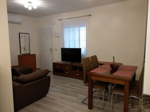 Ein Wohnzimmer mit einer Couch, einem Fernseher, einem Esstisch mit Stühlen und einem Fenster mit Vorhängen.