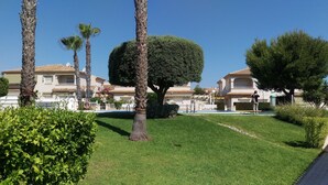 A well-maintained lawn with a neatly trimmed topiary tree, palm trees, and a row of Mediterranean-style villas with balconies and tiled roofs.