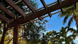 Una bottiglia di vino e un bicchiere su un tavolo sotto una pergola, con vista su alberi e un lanterna.