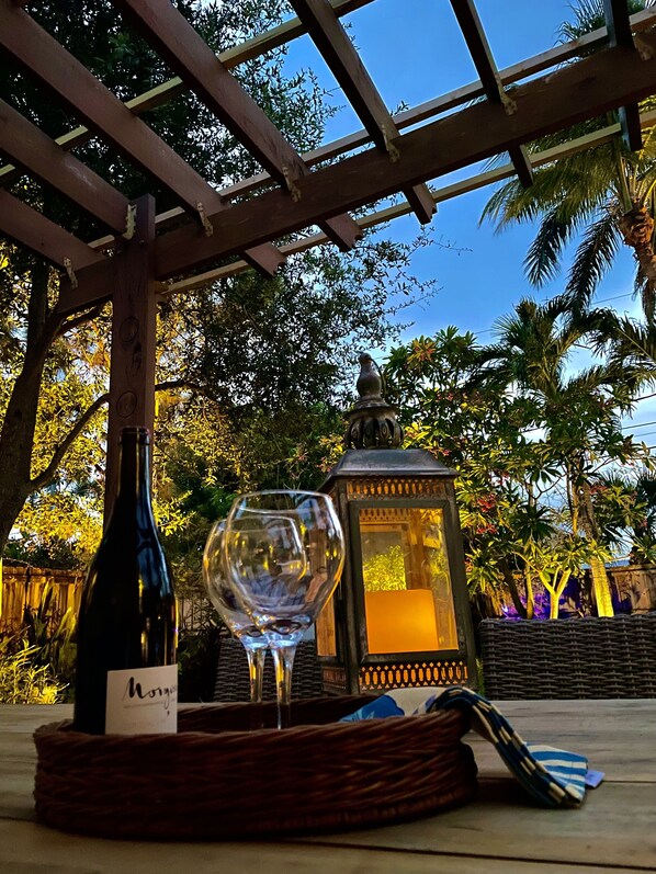 Une bouteille de vin et un verre sur une table placée sous une pergola, avec vue sur des arbres et une lanterne.