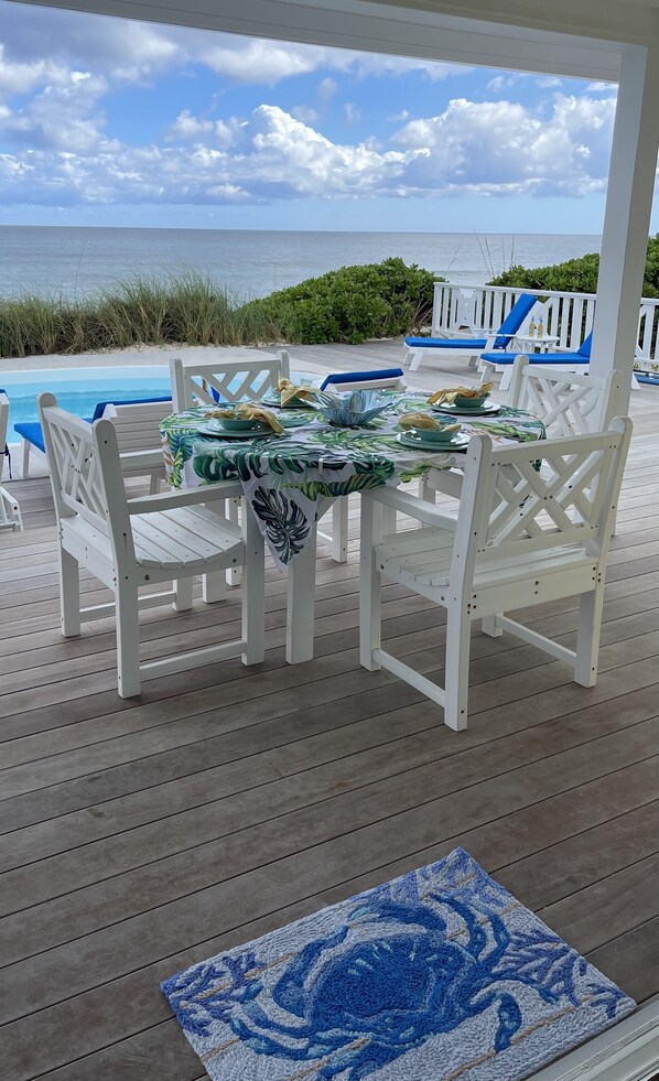 A covered outdoor dining area with white wooden chairs and a table set for two, overlooking a pool and beach.