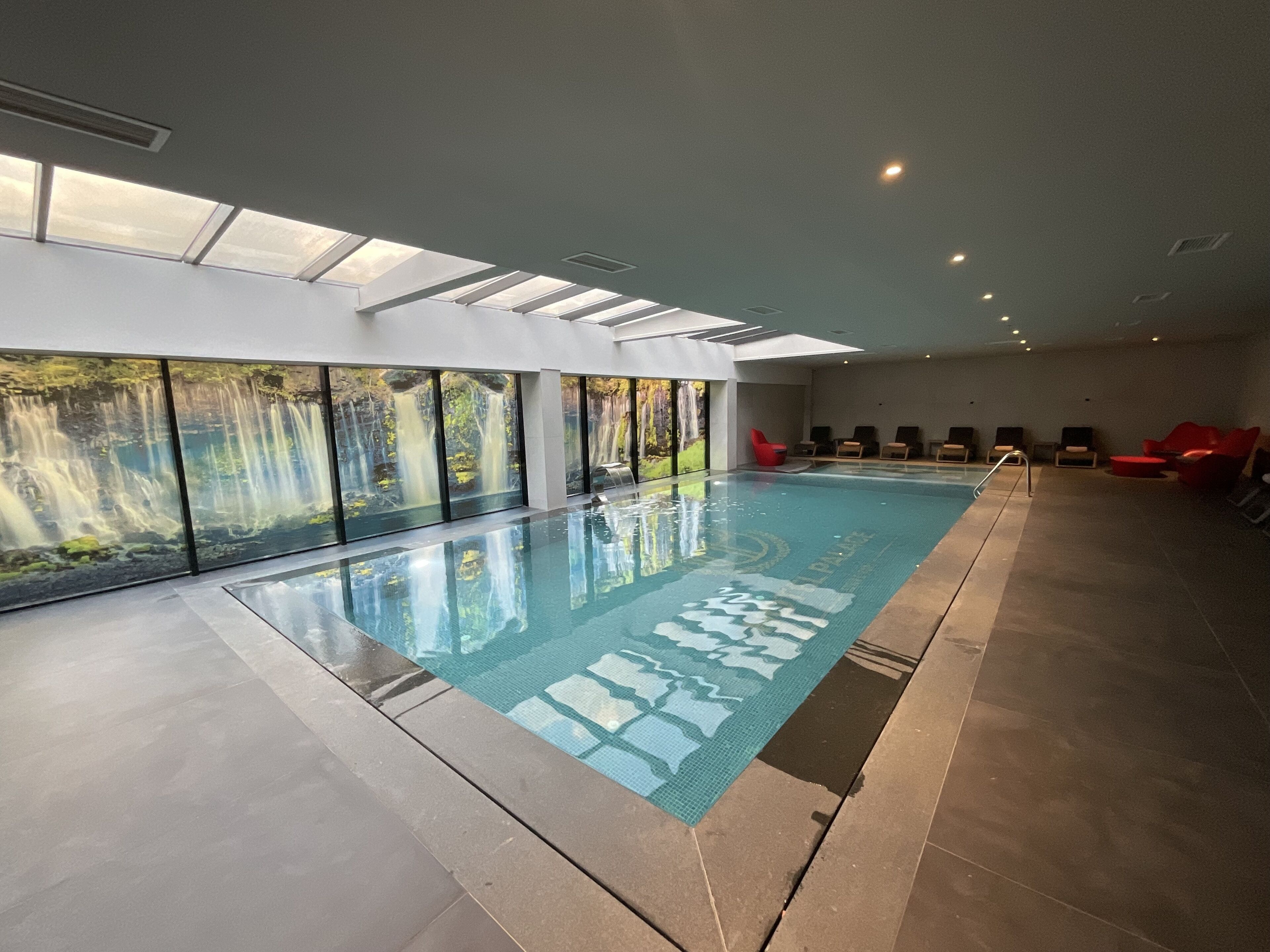 A large indoor swimming pool with clear glass walls, red chairs, and a waterfall mural.