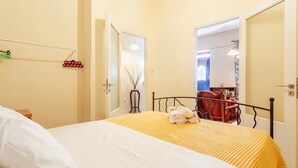 A hotel room with a bed, a chair, and a table with a lamp. There is a door leading to another room.