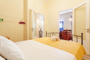 A hotel room with a bed, a chair, and a table with a lamp. There is a door leading to another room.