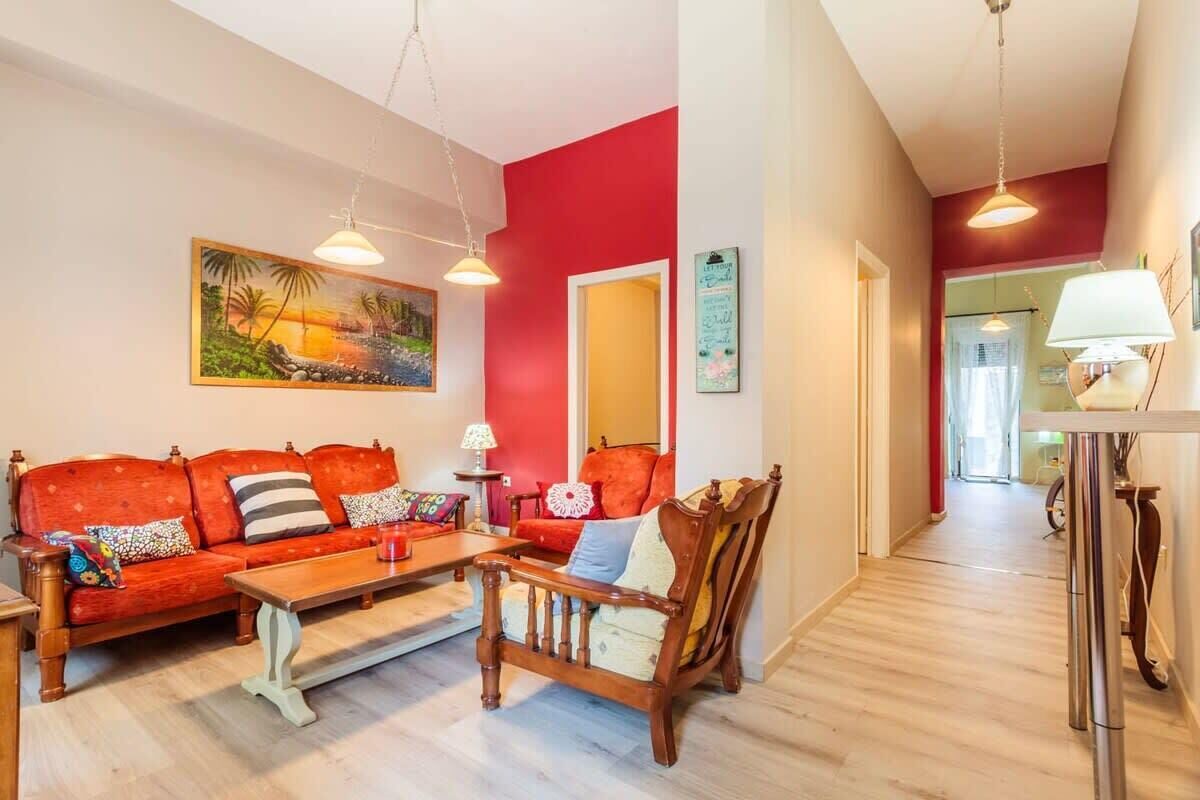 Un salon avec un mur rouge, un canapé en bois, un fauteuil en bois, une table basse et un tableau représentant une scène de plage.