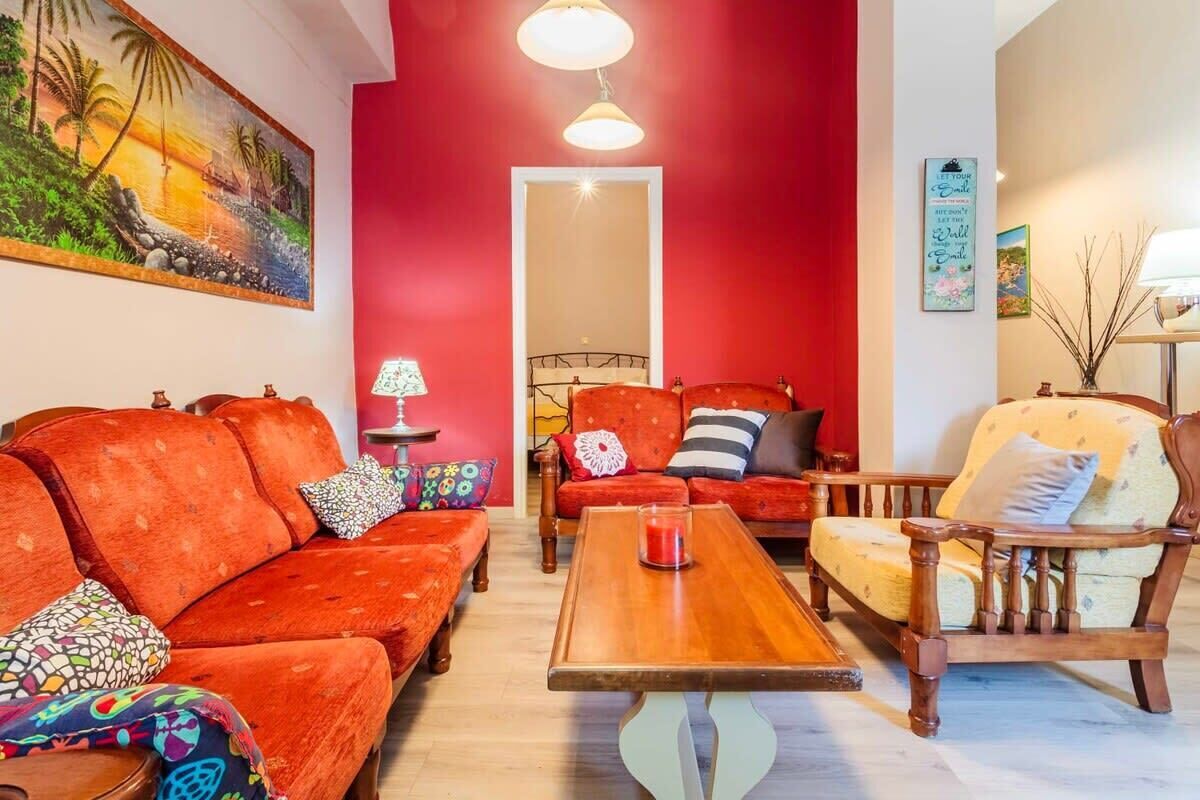 Ein Zimmer mit roter Wand, einem Holztisch, einer Couch, einem Sessel und einem Gemälde mit Strandmotiv.