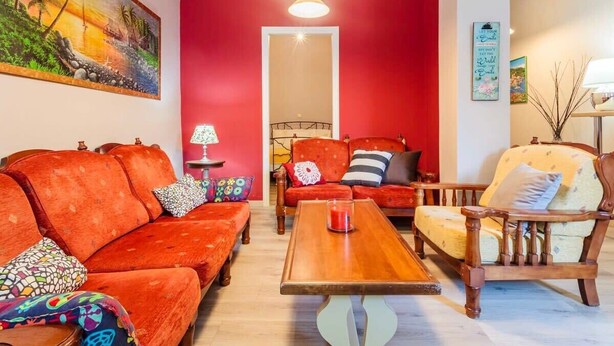 A room with a red wall, a wooden coffee table, a sofa, a chair, and a painting of a beach scene.