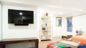 Quarto com televisão na parede, um banquinho de madeira, estante com objetos decorativos, cama com manta laranja e janela com persianas.