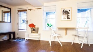 Quarto com piso de madeira, paredes brancas, um banco branco, uma cadeira branca, uma mesa pequena com um cesto e um quadro na parede.