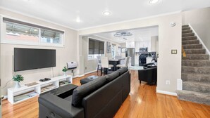Een moderne woonkamer met een flatscreen televisie, een zwarte leren bank, houten vloeren en een keuken op de achtergrond.