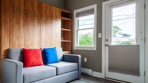 Un sofá gris con cojines rojos y azules, un mueble de madera, una ventana y una puerta que ofrece vistas a zonas verdes y edificios.