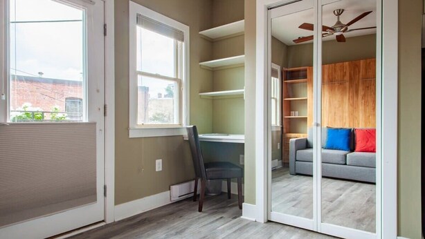 Una habitación con puerta corrediza que da a un balcón, un escritorio con silla, un sofá gris con cojines de colores y un ventilador de techo.