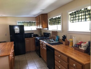 Una cocina con armarios de madera, un refrigerador negro, una estufa y un lavavajillas. Hay ventanas con cortinas verde y blanco, y un ventanal con cortina de cuadros.