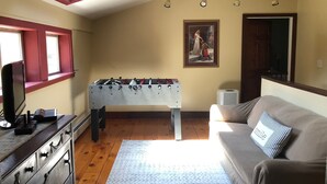 Una sala de estar acogedora con una mesa de fútbol de mesa, un sofá con un cojín con forma de sonrisa, un televisor y un cuadro en la pared.