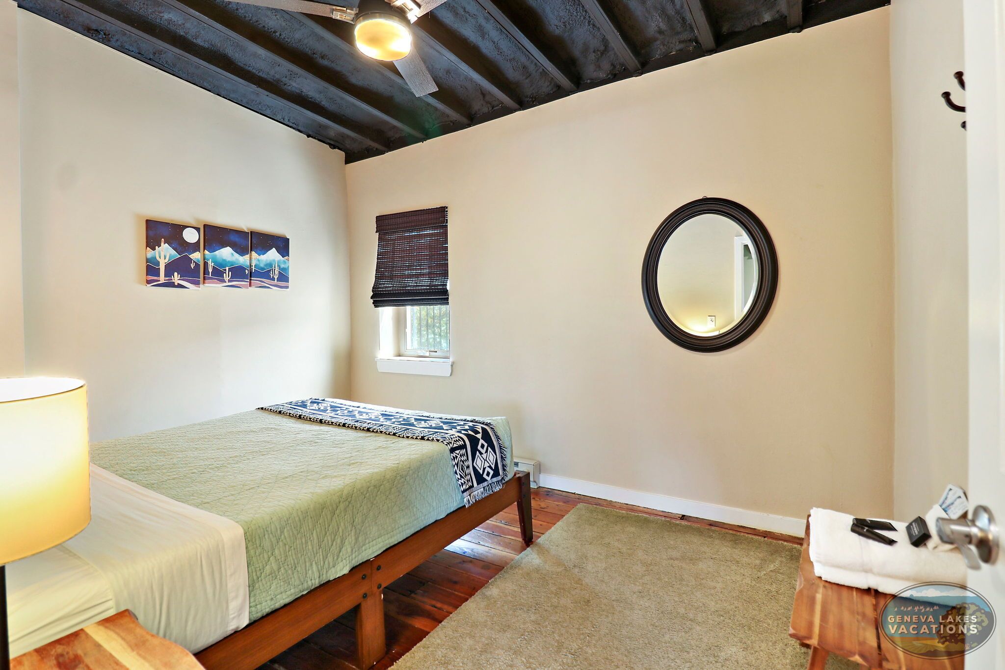 A bedroom with a bed, a bedside lamp, a window with blinds, a mirror, and a small table with a towel and toiletries.