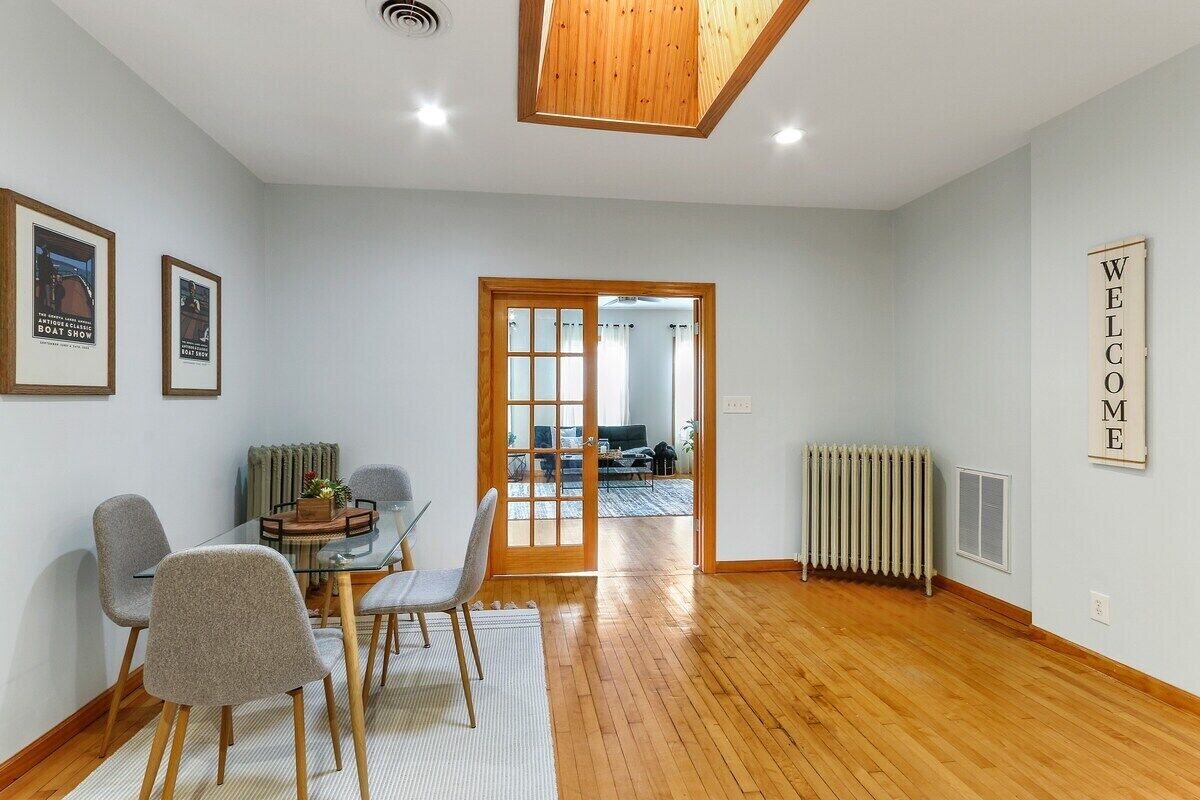 Una sala de estar moderna con una mesa de comedor de vidrio, sillas tapizadas en gris, piso de madera, un radiador y un letrero en la pared que dice 'WELCOME'.