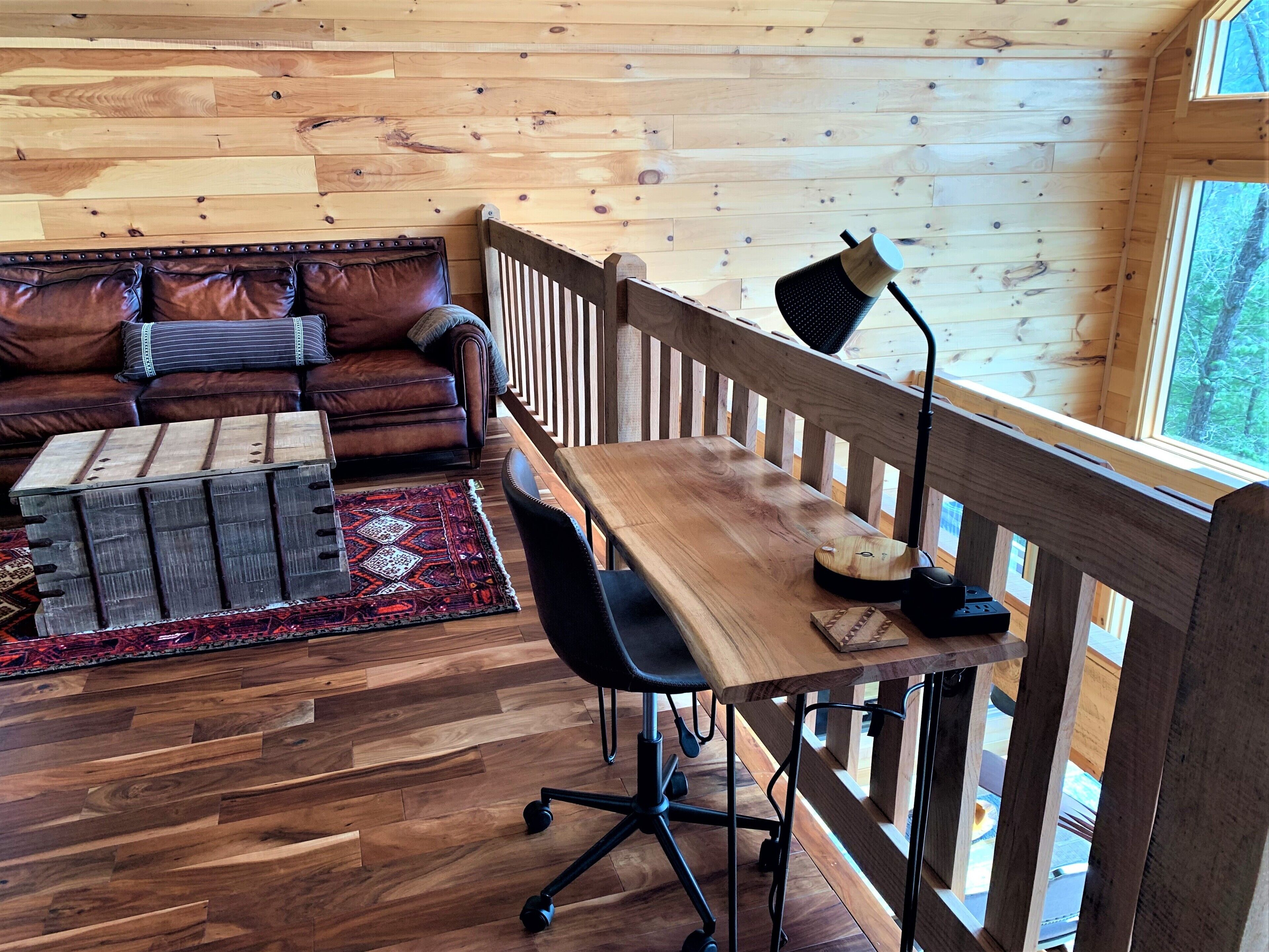 Una habitación acogedora con piso de madera, un sofá de cuero, un escritorio de madera con una silla y un ventanal grande.