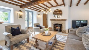 A living room with exposed wooden beams, a fireplace, a television, and patterned furniture. - A luxury holiday cottage in the Cotswolds - Manor Farm Cottage (Donnington, near Stow-on-the-Wold)