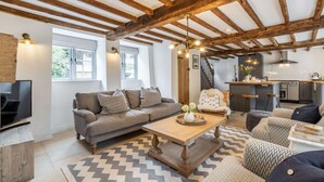 A cozy living room with exposed wooden beams, a sofa set, a coffee table, and a kitchen area visible through an open door. - A luxury holiday cottage in the Cotswolds - Manor Farm Cottage (Donnington, near Stow-on-the-Wold)