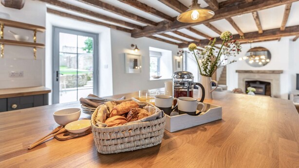 A kitchen with a wooden table, a basket of bread, a coffee maker, and a vase with flowers. - A luxury holiday cottage in the Cotswolds - Manor Farm Cottage (Donnington, near Stow-on-the-Wold)