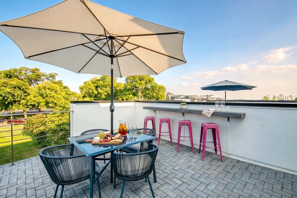 A rooftop terrace with a table set for dining, two pink bar stools, a large umbrella, and a city view.