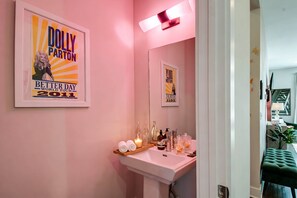 Une salle de bain avec un mur rose, une affiche encadrée de Dolly Parton, un lavabo avec des articles de toilette et un miroir.