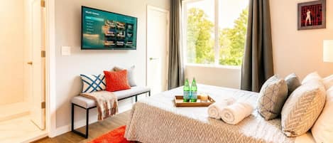 A hotel room with a bed, a TV mounted on the wall, a window with curtains, and a small table with bottles and a candle.