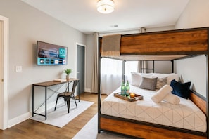 A bunk bed with a wooden headboard, a desk with a chair, a TV mounted on the wall, and a window with curtains.