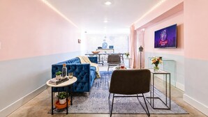 A modern hotel room with a blue sofa, a small round table with a plant, a black chair, a glass coffee table, and a TV on the wall.