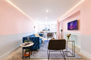A modern hotel room with a blue sofa, a small round table with a plant, a black chair, a glass coffee table, and a TV on the wall.