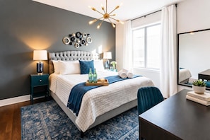 A modern bedroom with a tufted headboard, a large window with curtains, a bedside table with a lamp, a bedside plant, and a tray with bottles and a candle.
