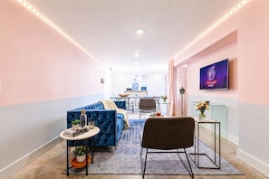 Una habitación de hotel moderna con un sofá azul, una pequeña mesa redonda con una planta, una silla negra, una mesa de centro de vidrio y un televisor en la pared.