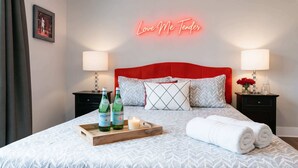 A neatly made bed with a red headboard, two bedside tables with lamps, a tray with bottles and a candle, and a neon sign above the bed.