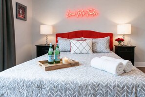 Un lit bien fait, avec une tête de lit rouge, deux tables de chevet avec des lampes, un plateau avec des bouteilles et une bougie, et un panneau lumineux au-dessus du lit.