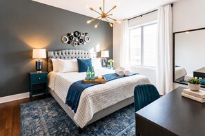 A modern bedroom with a tufted headboard, a large window with curtains, a bedside table with a lamp, a bedside plant, and a tray with bottles and a candle.