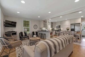 Ein modernes Wohnzimmer mit einer Couch, Sesseln und einem Couchtisch. Es gibt eine Küchenzeile mit Schränken, einer Mikrowelle und einem Kühlschrank.