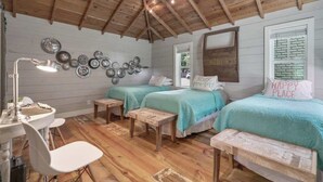 Quarto aconchegante com piso de madeira, duas camas com roupa de cama azul-turquesa, uma escrivaninha com luminária e um painel decorativo com placas de metal.