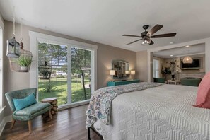 A bedroom with a large bed, a ceiling fan, a window with blinds, a chair, and a small table.