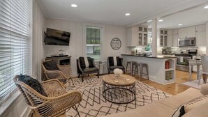 Uma sala de estar moderna com móveis de vime, uma mesa de centro redonda e uma cozinha com armários brancos e eletrodomésticos de aço inoxidável.
